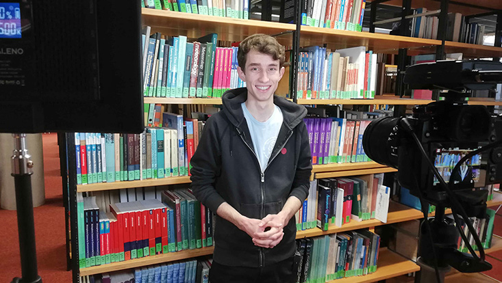 Ein junger Mann steht in einer Bibliothek und davor stehen eine Kamera und Lampen.
