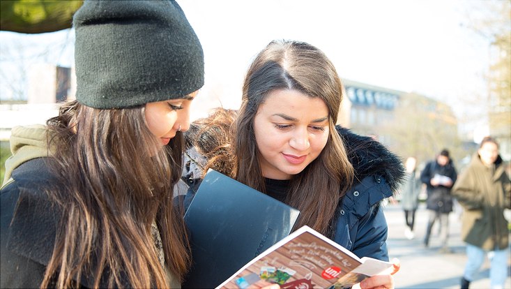 Zwei jung Frauen auf dem Campus, sie schauen in eine Informationsbroschüre der Universität